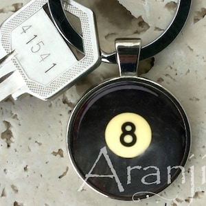 May include: A silver keychain with a black and white 8-ball charm. The charm has the number 8 in white on a yellow background. The text "Aranji" is printed on the charm.