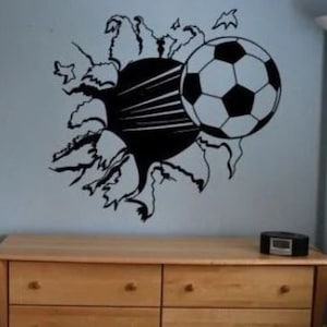 May include: Black and white soccer ball decal on a light blue wall, appearing as if it is breaking through the wall. The decal is above a wooden dresser with four drawers.