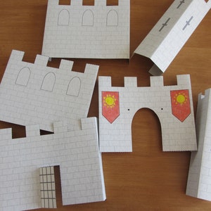 May include: A set of paper castle pieces for a craft project. The pieces are white with gray brick patterns and include a gate, towers, and a wall with red and yellow flags.