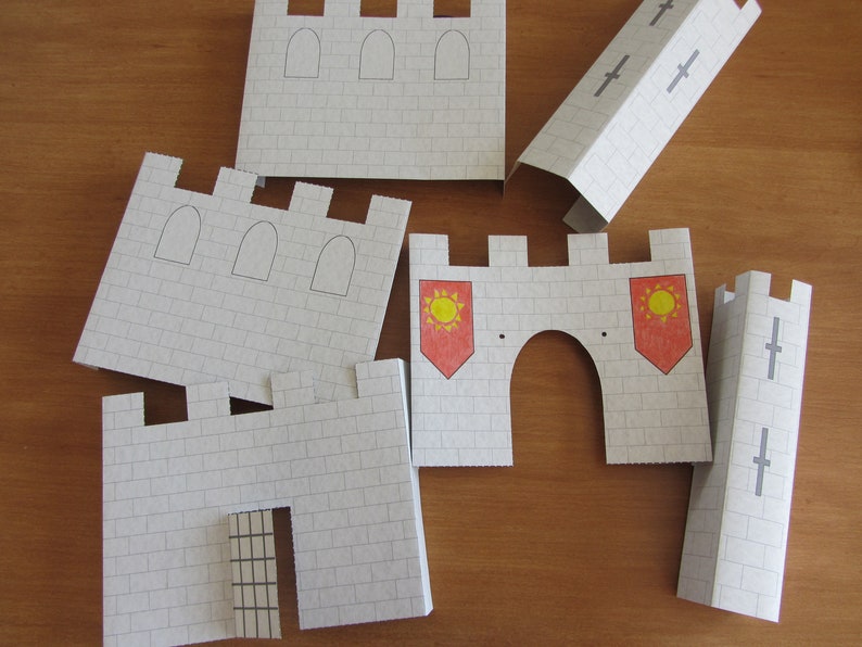 May include: A set of paper castle pieces for a craft project. The pieces are white with gray brick patterns and include a gate, towers, and a wall with red and yellow flags.