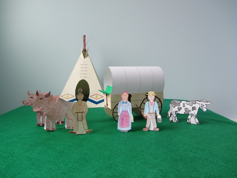 May include: A papercraft scene depicting a covered wagon, a teepee, and figures of a Native American, a woman, and a man. The wagon is pulled by two brown oxen, and the man is holding a rifle. The scene is set against a green background.