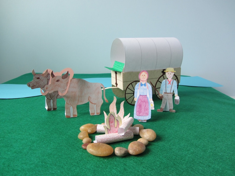 May include: A paper model of a covered wagon pulled by two oxen, with a couple of figures standing nearby. The wagon is white with a green roof and brown wheels. The oxen are brown with pink noses. The figures are wearing blue and brown clothing. There is a small campfire in front of the wagon.