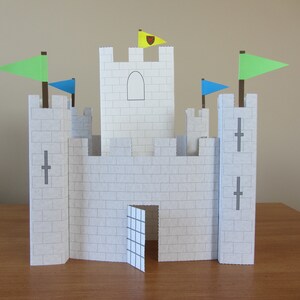 May include: A white paper castle with a gray door and gray crosses on the towers. The castle has three flags, one yellow with a red heart, one green, and one blue.