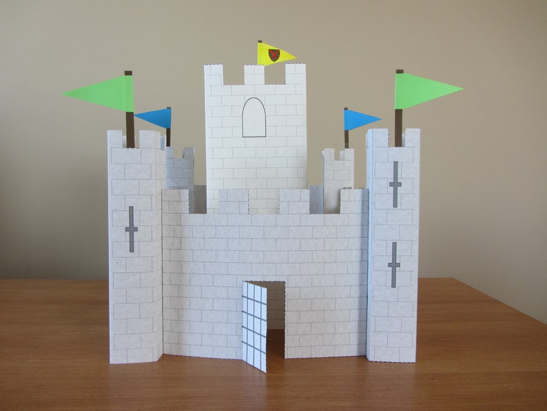 May include: A white paper castle with a gray door and gray crosses on the towers. The castle has three flags, one yellow with a red heart, one green, and one blue.
