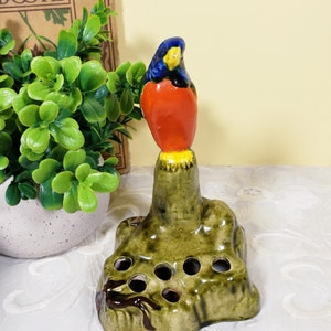 May include: A ceramic bird figurine with blue, red, and yellow paint sits atop a green ceramic base with six holes. The base is shaped like a tree stump.