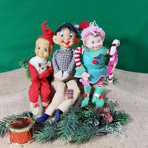 May include: Three vintage Christmas elves sitting on burlap fabric. The elf on the left is wearing a red and white outfit. The elf in the middle is wearing a black and white checkered shirt and tan pants. The elf on the right is wearing a green and white outfit with a pink and white hat and a candy cane.