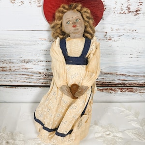 May include: Antique cloth doll with a red woven hat and a cream-colored dress with a floral pattern. The doll has curly blonde hair, blue eyes, and a painted face. The dress has navy blue trim and the doll is holding a heart-shaped object.