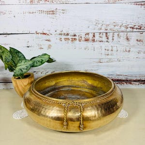 May include: A large, hammered brass bowl with a wide, shallow design. The bowl features a decorative rope-like trim around the rim and two rope accents. The interior has a golden hue, and the exterior shows a slightly aged patina. A decorative piece.