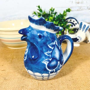 May include: A blue and white ceramic pitcher shaped like a rooster with a white and blue checkered pattern on the bottom. The rooster has a blue beak and eyes and is decorated with blue fruit and leaves.