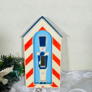 Puede incluir: Una cubierta decorativa para interruptor de luz con diseño de soldado. La cubierta tiene un techo azul, lados a rayas rojas y blancas, y una figura de soldado azul con una abertura de interruptor negra. El diseño recuerda a una garita.