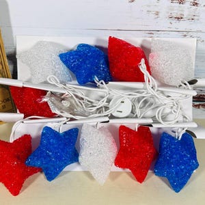 May include: A string of decorative lights featuring ten star-shaped covers in alternating red, white, and blue colors. The stars have a textured, crystalline appearance and are connected by a thin white electrical cord with a standard plug.
