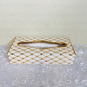 May include: A rectangular tissue box with a white background and a gold diamond pattern. The edges and the opening are trimmed with gold. The tissue box is sitting on a white lace tablecloth.