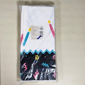 Puede incluir: Un mantel blanco con temática de cumpleaños, empaquetado en plástico transparente. El diseño incluye velas coloridas, un borde negro con formas geométricas y el texto "TABLE COVER". El mantel mide 137 cm por 259 cm.