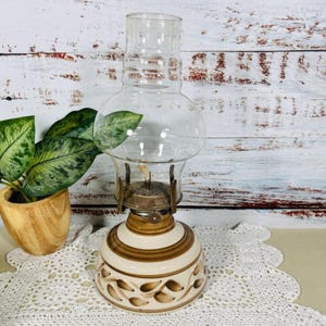 Puede incluir: Una lámpara de aceite vintage con un globo de vidrio transparente y una base de cerámica. La base es de color crema con rayas marrones y diseños en forma de hoja. La lámpara tiene un soporte de mecha de metal.