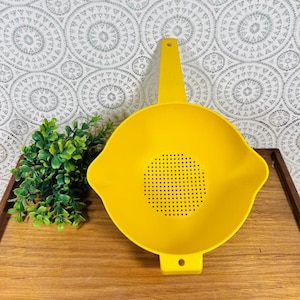 Puede incluir: Un colador amarillo brillante con un asa y una base perforada. Este utensilio de cocina de plástico está diseñado para escurrir líquidos de los alimentos. El colador está sobre una bandeja de madera.