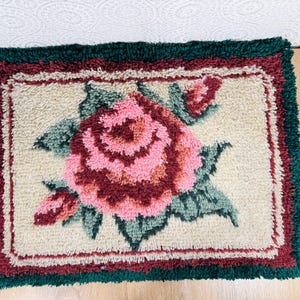 Könnte beinhalten: Ein rechteckiger Knüpfteppich mit einem zentralen rosa Rosenmotiv und grünen Blättern auf cremefarbenem Grund. Der Teppich ist von einer dekorativen Bordüre umrahmt, die aus einer dünnen kastanienbraunen Linie und einem dunkelgrünen Rand besteht.
