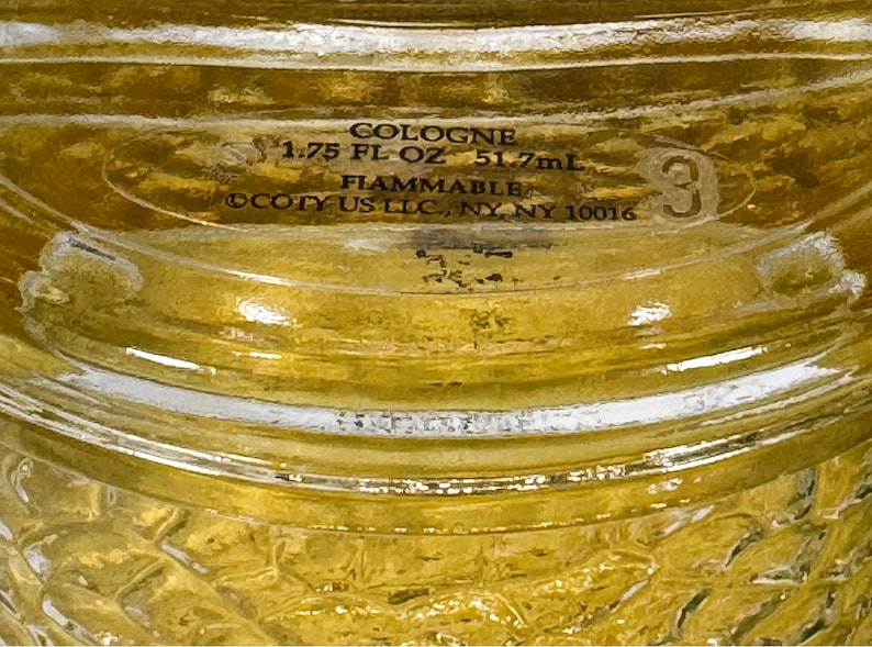 Puede incluir: Una botella de vidrio transparente con una etiqueta dorada que dice "COLOGNE 1.75 FL OZ 51.7mL FIAMMABLE COTY US LLC, NY, NY 10016". La botella tiene una superficie texturizada y un n&uacute;mero "3" impreso en el lateral.
