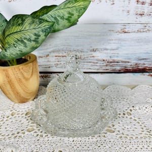 Puede incluir: Mantequillera de cristal tallado transparente con tapa abovedada y diseño de estrella. El plato se asienta sobre una base festoneada a juego. Un pequeño pomo decorativo remata la tapa. La mantequillera se exhibe sobre un tapete de encaje blanco.