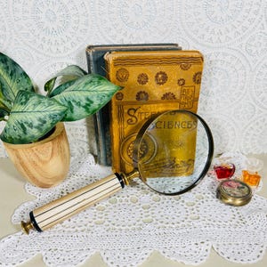 May include: A magnifying glass with a cream and black striped handle and a brass rim, positioned in front of two vintage books, one with a yellow cover titled Steele's Sciences, a small round floral tin, and two miniature glass perfume bottles.