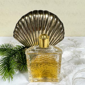 Puede incluir: Una botella de perfume de vidrio transparente con una tapa dorada, sentada sobre un soporte de concha de mar plateada. La botella est&aacute; llena de un l&iacute;quido dorado y tiene una superficie texturizada.
