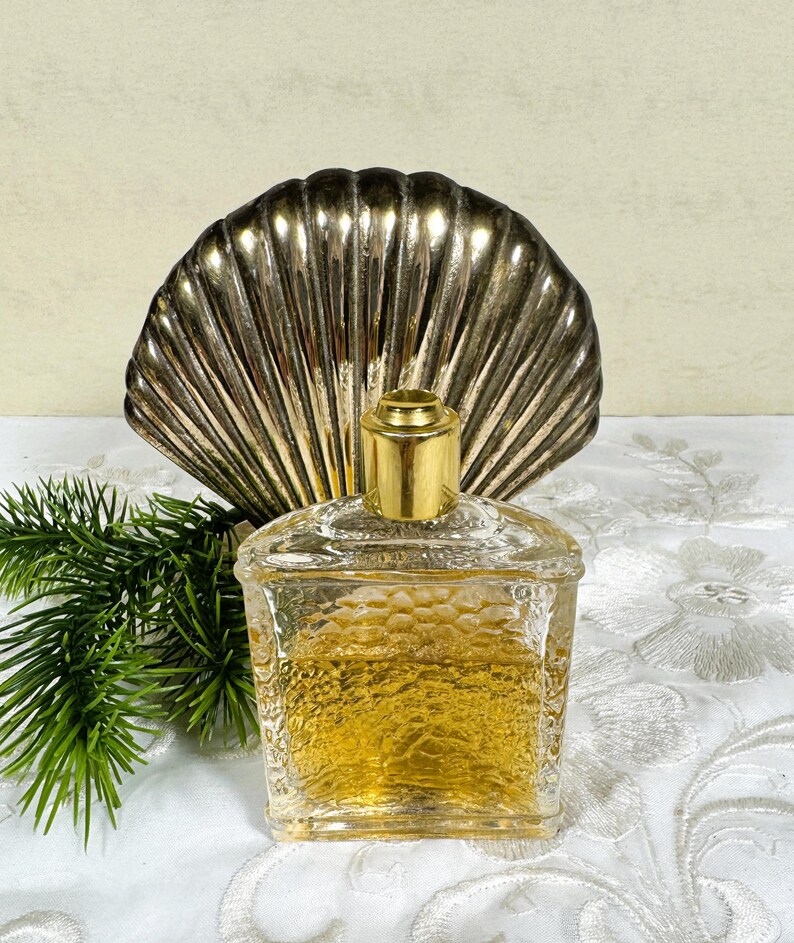 Puede incluir: Una botella de perfume de vidrio transparente con una tapa dorada, sentada sobre un soporte de concha de mar plateada. La botella est&aacute; llena de un l&iacute;quido dorado y tiene una superficie texturizada.