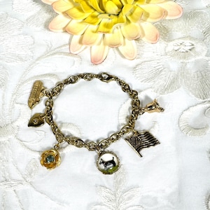 May include: A gold charm bracelet featuring a selection of charms, including a heart, a flower, a ship, a bell, and an American flag. The bracelet is displayed against a white background.