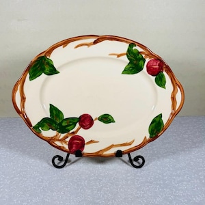 May include: Oval serving platter with a cream-colored surface, adorned with a brown branch design featuring red apples and green leaves. The rim has a scalloped edge. The platter is displayed on a black metal stand.