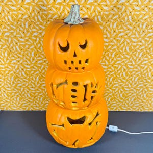May include: A stacked orange ceramic pumpkin decoration with carved-out facial features and bone designs. The top pumpkin has crescent-shaped eyes and a triangular nose. The stem is green. A white cord and switch are attached to the bottom.