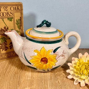 Puede incluir: Tetera de cerámica color crema con un diseño de girasol amarillo. La tapa tiene rayas verdes y amarillas y un asa verde. Un libro vintage está en el fondo.