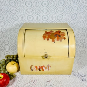 May include: A vintage, cream-colored bread box with a roll-top lid. The box features mushroom decals on the front and top. A small, decorative knob serves as the handle. The bread box is made of metal and has a slightly curved design.