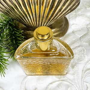 Puede incluir: Una botella de perfume de vidrio transparente con una tapa dorada y una superficie texturizada. La botella est&aacute; medio llena de un l&iacute;quido amarillo claro.