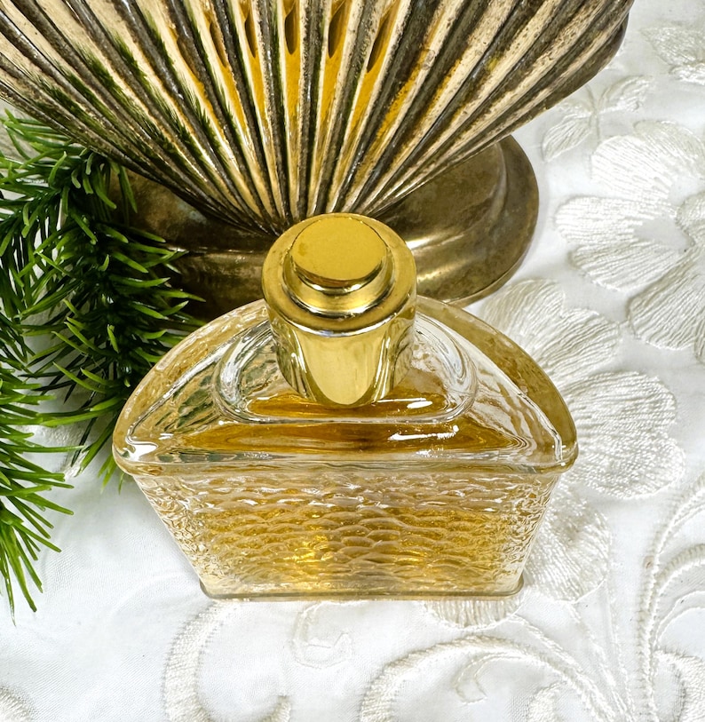 Puede incluir: Una botella de perfume de vidrio transparente con una tapa dorada y una superficie texturizada. La botella est&aacute; medio llena de un l&iacute;quido amarillo claro.