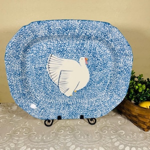 May include: A blue and white speckled ceramic serving platter with a white turkey illustration. The platter has a scalloped edge and is supported by a black metal stand. The turkey has a red beak and yellow feet.