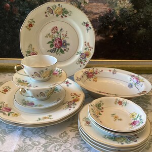 May include: A set of vintage floral china dinnerware with a white background and gold trim. The set includes a large dinner plate, a salad plate, a soup bowl, a teacup and saucer, and a set of dessert plates.