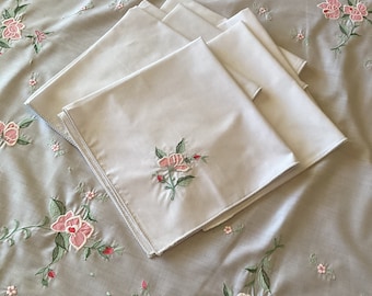 May include: Five white linen napkins with delicate pink floral embroidery. The napkins are folded and stacked on a light gray background with a matching floral pattern.
