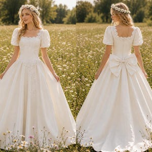 Op de afbeelding: Een witte trouwjurk met een vierkante halslijn, korte pofmouwen en een volle rok. De voorkant van de jurk heeft delicate kanten details, terwijl de achterkant een grote strik heeft. De bruid draagt een bloemenkrans.
