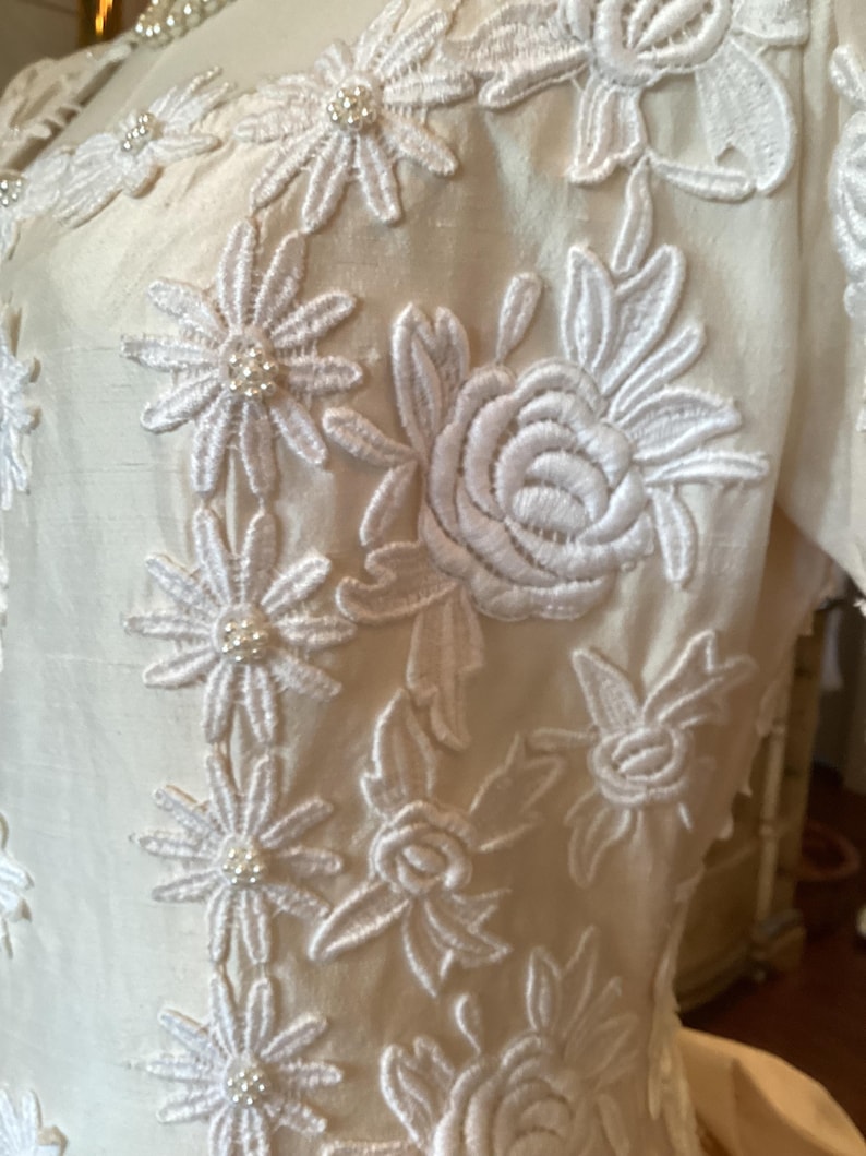 Op de afbeelding: Ivoor kleurige jurk met ingewikkelde witte bloemenborduurwerk. Het borduurwerk bevat madeliefachtige bloemen en rozen, met kleine parelaccenten. De stof heeft een subtiele glans, wat wijst op zijde of satijn.