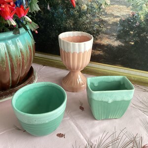 May include: A collection of ceramic planters in various shapes and colors. Includes a large turquoise and brown planter, a pink and white goblet-shaped planter, a green bowl, and a square green planter with a white rim.