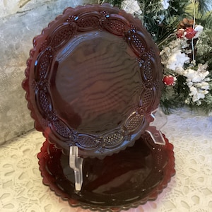 May include: Two ruby red glass plates with a scalloped edge and embossed grape and floral design. The plates are displayed on a clear plastic stand. The plates are likely vintage or antique and are suitable for decorative use.