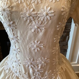 Op de afbeelding: Cr&egrave;mekleurige vintage jurk met bloemenborduurwerk. De jurk heeft een vierkante halslijn en korte mouwen. Witte madeliefachtige geborduurde bloemen met parelaccenten sieren het lijfje en de rok. De rok heeft een volle, vloeiende vorm, waardoor een klassiek silhouet ontstaat.