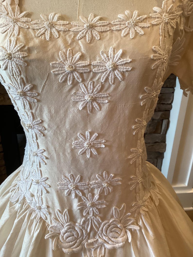 Op de afbeelding: Cr&egrave;mekleurige vintage jurk met bloemenborduurwerk. De jurk heeft een vierkante halslijn en korte mouwen. Witte madeliefachtige geborduurde bloemen met parelaccenten sieren het lijfje en de rok. De rok heeft een volle, vloeiende vorm, waardoor een klassiek silhouet ontstaat.