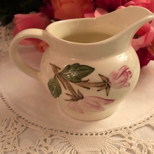 May include: A white ceramic creamer with a floral design featuring pink roses and green leaves. The creamer is sitting on a white crocheted doily.