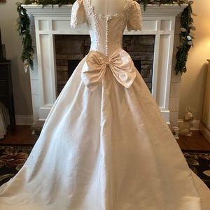 Op de afbeelding: Een vintage ivoorkleurige trouwjurk met een grote strik in de taille. De jurk heeft een getailleerd lijfje met delicate kanten details en korte mouwen. De volle rok valt tot op de grond en cre&euml;ert een klassiek en elegant silhouet.