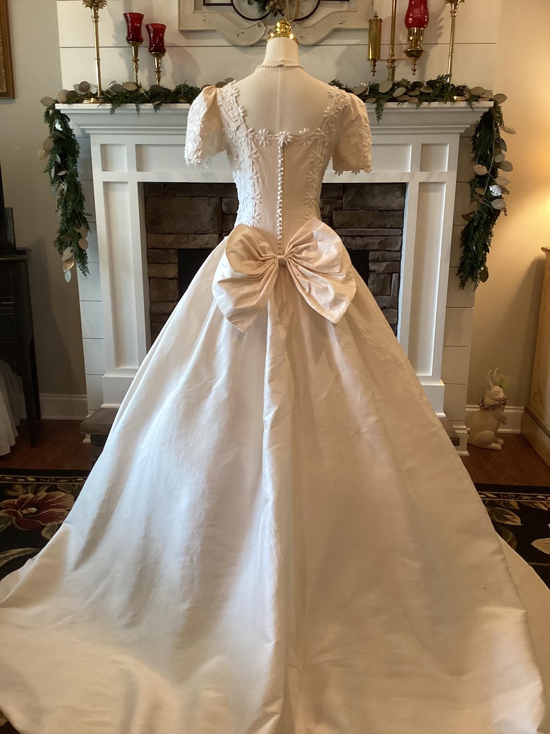 Op de afbeelding: Een vintage ivoorkleurige trouwjurk met een grote strik in de taille. De jurk heeft een getailleerd lijfje met delicate kanten details en korte mouwen. De volle rok valt tot op de grond en cre&euml;ert een klassiek en elegant silhouet.