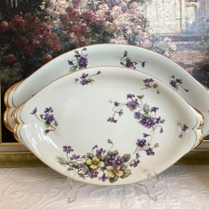May include: Two white oval serving platters with gold trim and a floral design featuring purple and yellow flowers. The platters have scalloped edges.