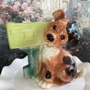 May include: A ceramic figurine of a brown and black spotted dog holding a green mailbox. The dog is sitting on a white surface.