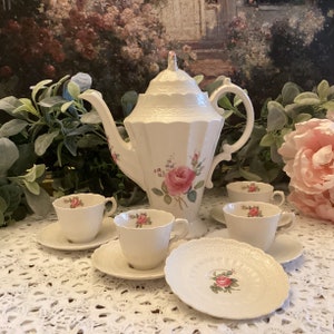 May include: A white porcelain teapot with a floral design and four matching teacups and saucers. The teapot has a lid with a scalloped edge and a handle on the side. The teacups and saucers have a scalloped edge and a floral design. The tea set is on a white crocheted tablecloth.
