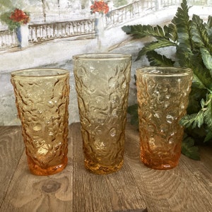 May include: Three amber glass drinking glasses with a textured, bubbled surface. The glasses are different sizes and are arranged in a row on a wooden surface.