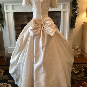 Op de afbeelding: Een vintage ivoorkleurige trouwjurk op een mannequin. De jurk heeft een getailleerd lijfje met bloemenkant, korte mouwen en een grote strik in de taille. De volle rok valt tot op de grond. De achterkant van de jurk heeft een rij knopen.