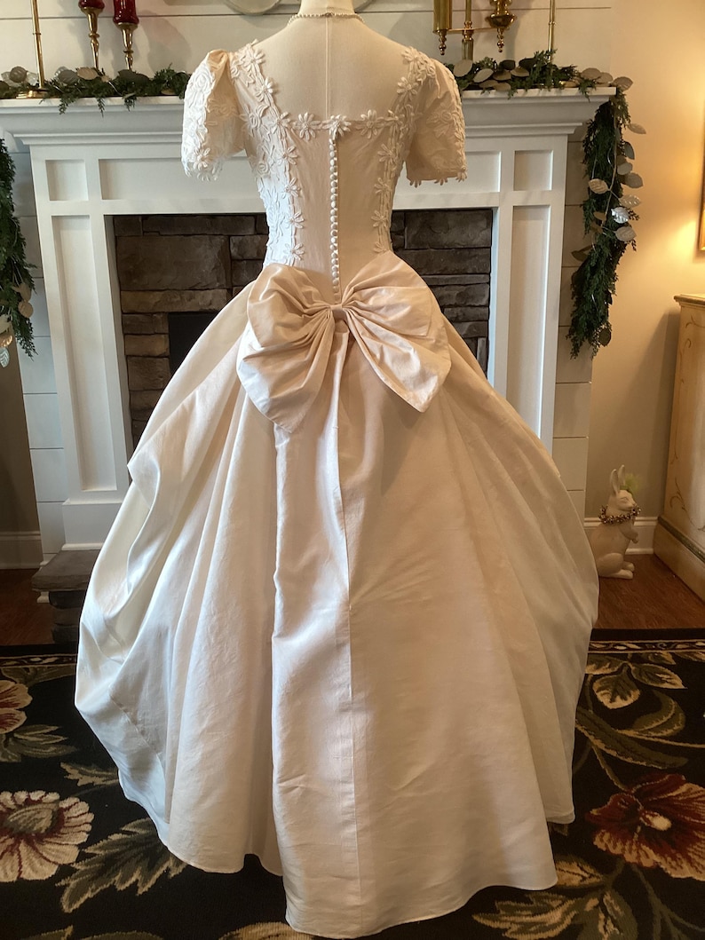 Op de afbeelding: Een vintage ivoorkleurige trouwjurk op een mannequin. De jurk heeft een getailleerd lijfje met bloemenkant, korte mouwen en een grote strik in de taille. De volle rok valt tot op de grond. De achterkant van de jurk heeft een rij knopen.